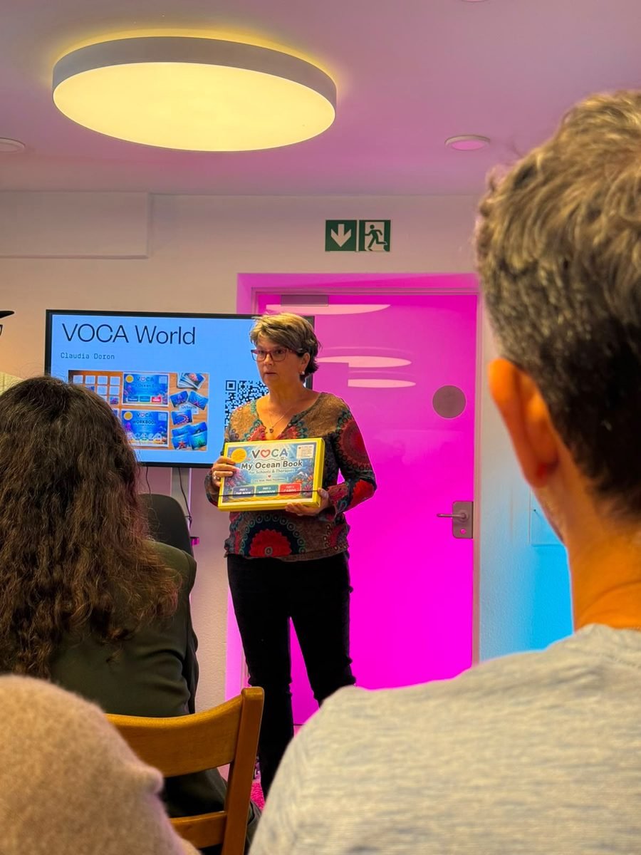Claudia Doron steht vor einer Leinwand mit der Aufschrift VOCA World und hält das VOCA-Buch hoch.
